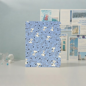 Può includere: Un taccuino rettangolare con una copertina azzurra caratterizzata da un motivo ripetuto di Snoopy in varie pose. Le illustrazioni a linee bianche e nere del personaggio sono sparse su tutta la superficie della copertina anteriore.
