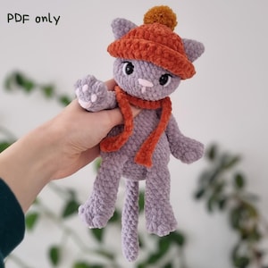 Könnte beinhalten: Gehäkelte Katzendoll in Lavendel mit orangefarbener Mütze und Schal. Die Katze hat schwarze Augen und eine rosa Nase. Der Text "PDF only" befindet sich oben links. Handgefertigt. Ein charmantes Produkt.