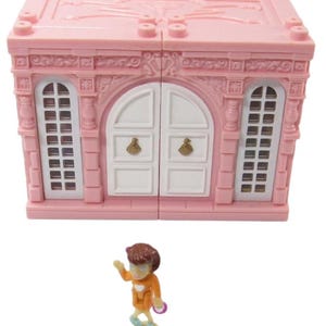May include: A pink plastic toy house with white doors and windows. A small plastic doll with brown hair and an orange dress is standing in front of the house.