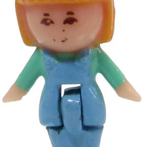 May include: A small plastic doll with orange hair, wearing a teal shirt and blue overalls. The doll has a light skin tone and is standing with arms outstretched. The doll's shoes are green.