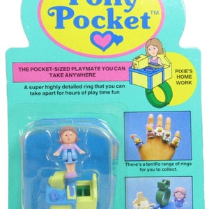 May include: A Polly Pocket ring playset featuring a blue and yellow desk and a small doll. The packaging is blue and yellow with the text "Polly Pocket" and "Pixie's Home Work".