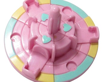 May include: A pink and pastel-colored toy with a circular design. The toy features a central column, heart-shaped accents, and segmented sections in pink, yellow, and teal. The toy has a playful and colorful appearance.
