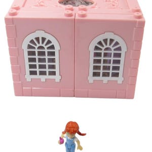 May include: A pink plastic toy house with two windows and a white roof. A small red-haired doll with a blue dress and a pink flower is standing in front of the house.