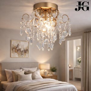 May include: A gold-toned, flush-mount chandelier with cascading crystal accents. The fixture features a central light source surrounded by ornate metalwork and hanging crystal prisms. The chandelier is installed in a bedroom setting, adding a touch of elegance.