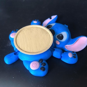 Peut inclure: Figurine bleue imprimée en 3D d'un personnage de dessin animé avec de grandes oreilles, des yeux noirs et des accents roses. Un dessous de verre rond de couleur beige est placé sur le ventre du personnage. La figurine est sur une surface noire.