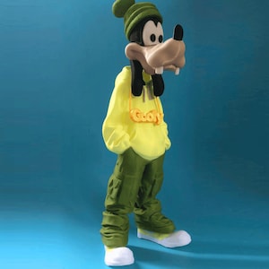 Puede incluir: Una figura de Goofy con una sudadera con capucha amarilla, pantalones cargo verdes y un gorro verde. La figura tiene un collar dorado que dice "Goofy". Los zapatos son blancos y verdes. El fondo es azul liso.