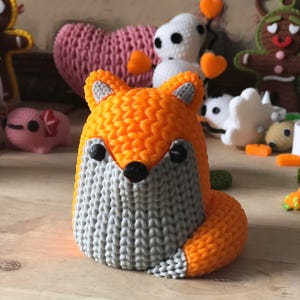 May include: A handcrafted orange and gray fox figurine with black eyes and nose. The fox has a textured, knitted appearance and is part of a collection of other handmade figurines, including a pink heart and a snowman.