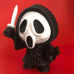 Puede incluir: Figura impresa en 3D del personaje Ghostface de la serie de películas de terror Scream. La figura es negra y blanca, sosteniendo un cuchillo blanco. Tiene una túnica con capucha negra y una máscara blanca.