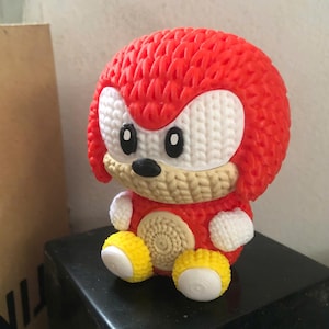 May include: A 3D-printed figurine of Knuckles the Echidna, a character from the Sonic the Hedgehog franchise. The figurine features a red body, white accents, and yellow shoes. The figure is sitting on a black surface.