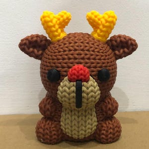 May include: A brown knitted reindeer figurine with yellow antlers, black eyes, and a red nose. The reindeer has a beige and black knitted design on its face. The figurine is a cute, handmade decoration.