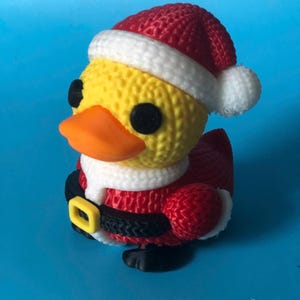 May include: A yellow duck figurine wearing a Santa Claus outfit, including a red knit hat with white trim, a white collar, and a black belt with a yellow buckle. The duck has an orange beak and black eyes. The background is a solid blue.