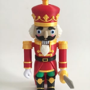 Puede incluir: Figura decorativa de cascanueces con diseño de soldado clásico. La figura presenta un abrigo rojo, detalles dorados y un bigote negro. Tiene barba blanca, pantalones verdes y botas negras. El cascanueces sostiene una espada plateada y lleva una corona roja.