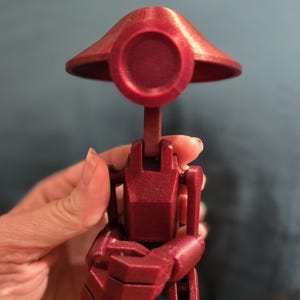 May include: A maroon-colored, articulated robot figurine with a circular head and a wide brim. The robot has a slender neck, a rectangular torso, and jointed arms and legs. The figurine is held in a hand.