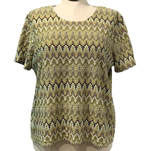 May include: A short-sleeved top with a round neckline. The shirt features a repeating leaf pattern in shades of green, brown, and cream. The fabric appears to have a textured surface. The top is displayed on a mannequin.