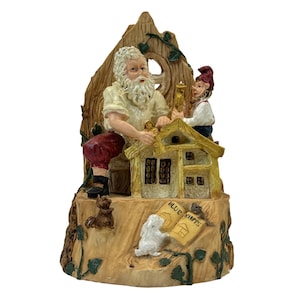 May include: A whimsical figurine depicting a bearded man and a gnome building a miniature house. The figurine is made of painted resin and features a detailed design with a tree-like backdrop, small animals, and blueprints. The text "BLUE PRINTS" is visible.