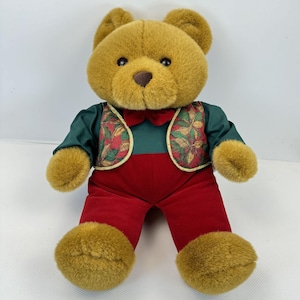 Könnte beinhalten: Ein Teddybär aus Plüsch mit goldbraunem Fell. Der Bär trägt ein grünes Hemd, eine rote Hose und eine Weste mit floralem Muster. Eine rote Fliege vervollständigt das Outfit. Der Bär sitzt aufrecht.
