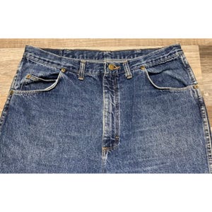 May include: Close-up of blue denim jeans. The jeans have a classic five-pocket design, a button and zipper closure, and belt loops. The denim has a faded appearance.
