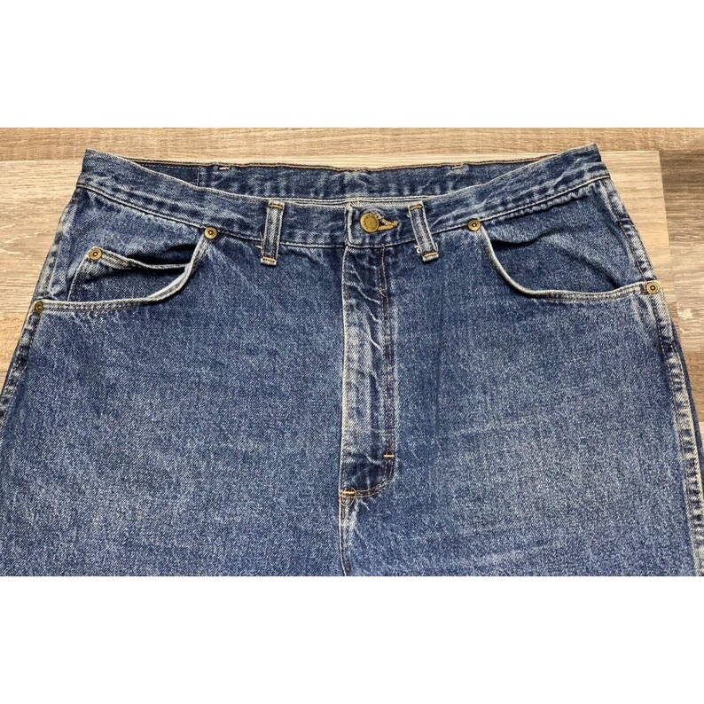 May include: Close-up of blue denim jeans. The jeans have a classic five-pocket design, a button and zipper closure, and belt loops. The denim has a faded appearance.