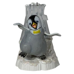 Könnte beinhalten: Graues Pinguin-Ornament mit schwarzem Kopf, gelber Fliege und ausgestreckten Flügeln. Der Pinguin steht auf einem weißen, glitzernden Sockel mit einer durchsichtigen Eisstruktur dahinter. Oben befindet sich eine silberne Schlaufe zum Aufhängen.