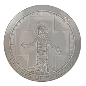 May include: A silver-colored round medallion featuring a depiction of a baby with outstretched arms, surrounded by a cross and a star. The text "19-KING OF KINGS-70" is inscribed around the edge of the medallion.
