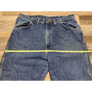 May include: Blue denim jeans with a straight leg cut. A yellow measuring tape is laid across the waist of the jeans, measuring approximately 24 inches across. The jeans have classic five-pocket styling.