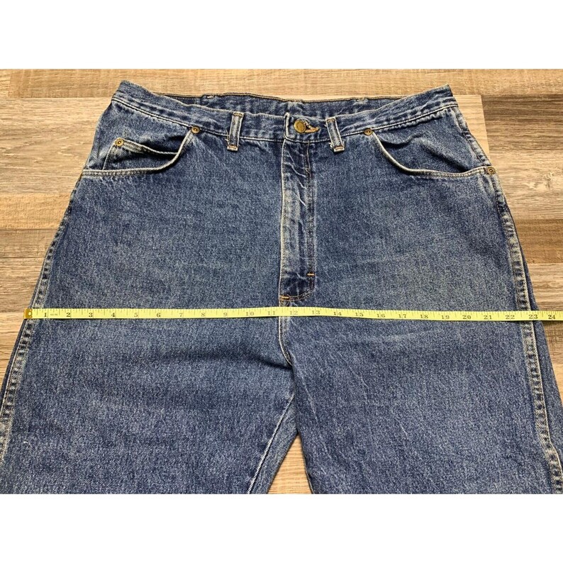 May include: Blue denim jeans with a straight leg cut. A yellow measuring tape is laid across the waist of the jeans, measuring approximately 24 inches across. The jeans have classic five-pocket styling.