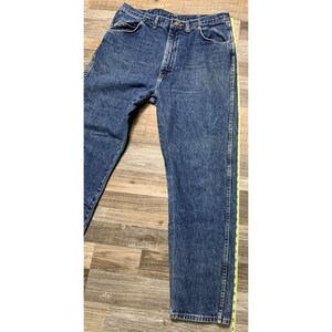 May include: Blue denim jeans with a classic five-pocket design. The jeans have a straight leg cut and a button and zipper closure. A yellow measuring tape is placed along the side of the jeans. The jeans are displayed on a wooden surface.