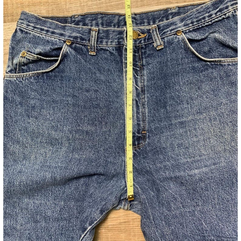 May include: Close-up of blue denim jeans with a measuring tape. The jeans have a classic five-pocket design, a button and zipper closure, and belt loops. The measuring tape shows a length of approximately 15 inches.