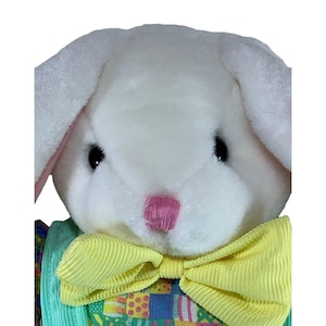 May include: A plush white bunny with black eyes and a pink nose. The bunny wears a yellow bow tie and a green vest with a colorful pattern. The bunny's ears have a pink interior.