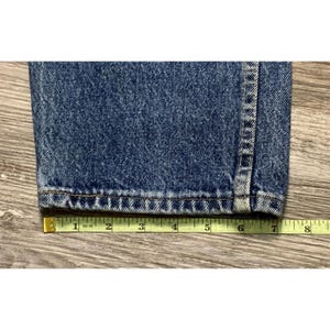 May include: Close-up of the hem of blue denim jeans. The jeans are measured with a yellow tape measure. The hem is approximately 7 inches wide.