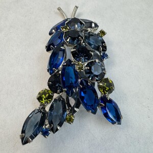 Könnte beinhalten: Vintage Brosche in Form einer Traube. Die Brosche hat eine silberfarbene Metalleinfassung und ist mit blauen und grünen Strasssteinen in verschiedenen Formen und Größen besetzt, die einen funkelnden Effekt erzeugen.