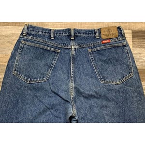May include: Close-up of the back of blue denim Wrangler jeans. The jeans have two back pockets, a brown leather patch with the Wrangler logo, and a small red tag. The denim is a medium wash.