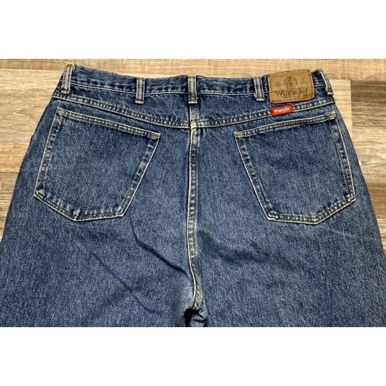 May include: Close-up of the back of blue denim Wrangler jeans. The jeans have two back pockets, a brown leather patch with the Wrangler logo, and a small red tag. The denim is a medium wash.