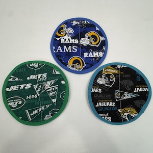 May include: Three circular fabric coasters featuring NFL team logos. One coaster displays the Los Angeles Rams logo in blue and yellow. Another shows the New York Jets logo in green and white. The third features the Jacksonville Jaguars logo in black and teal.