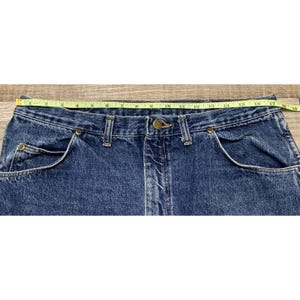 May include: Close-up of a pair of blue denim jeans with a yellow measuring tape across the waist. The jeans have classic five-pocket styling, a button and zipper closure, and measure approximately 18 inches across the waist.