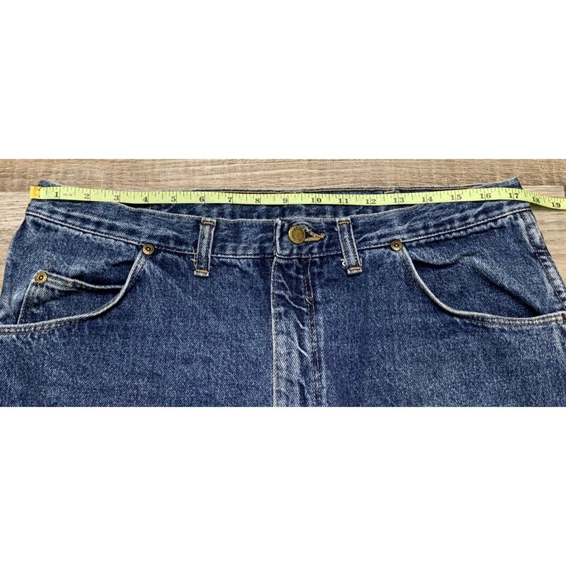 May include: Close-up of a pair of blue denim jeans with a yellow measuring tape across the waist. The jeans have classic five-pocket styling, a button and zipper closure, and measure approximately 18 inches across the waist.