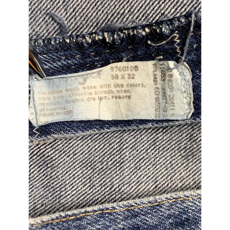 May include: Close-up of blue denim jeans with a tag. The tag displays the size 50 x 32 and washing instructions. The jeans have a classic five-pocket design and a button and zipper closure.