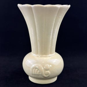 以下が含まれることがあります： 光沢のある仕上げのクリーム色の陶器製花瓶。広いスカラップ状の縁と、首元に沿った縦の溝模様が特徴です。丸みを帯びたベースの前面には、浮き彫りになった渦巻き状の装飾パターンが施されています。