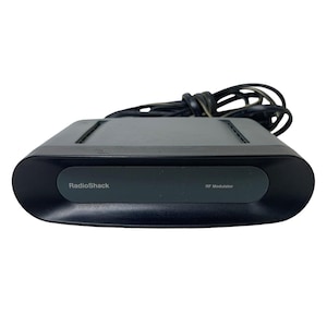 May include: A black RadioShack RF Modulator device with a smooth, rounded front panel and a dark gray center inlay. The unit features a black power cord coiled behind the main housing, which has ventilation slots along the top surface.