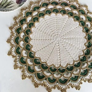 May include: A circular, handmade crochet doily. The centre is white, and the scalloped border features green, white, and gold sections. This decorative item could be used as a coaster or as a decorative piece for a table.