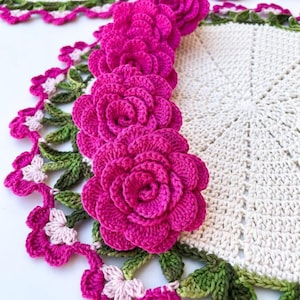 May include: A handmade crochet doily with a floral design. The doily has a cream-coloured centre with a border of pink and green crochet flowers and leaves. Several large, vibrant pink crochet roses are also present.