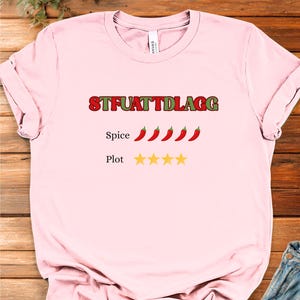 Puede incluir: Camiseta rosa claro con el texto "STFUATTDLAGG" en rojo y verde. Debajo, cuatro chiles rojos y cuatro estrellas amarillas. La camiseta está hecha de un material suave.