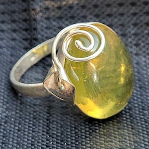 Peut inclure: Bague en argent ornée d'une grosse pierre ovale jaune-vert translucide. La pierre est sertie dans un anneau en argent avec un motif en spirale décoratif au-dessus. La bague a un style classique et élégant.