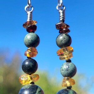 May include: Dangling earrings with a silver hook, featuring a combination of dark green and black marbled beads, amber-colored chips, and a dark red bead. The earrings are displayed against a bright blue sky.
