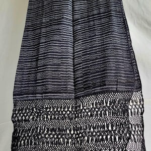 May include: A long rectangular black scarf featuring a fine white horizontal pinstripe pattern throughout the main body. The bottom edge is finished with an intricate, wide macrame-style knotted lattice border that transitions into long fringe.