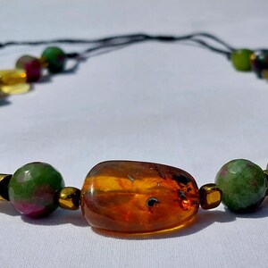 Peut inclure: Collier de perles avec une perle centrale ambrée. Il comprend des perles vertes, jaunes et rouges, avec des entretoises dorées. Le collier est monté sur un cordon noir.