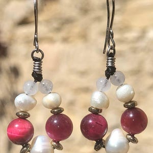 May include: A pair of handcrafted earrings featuring a circular design. The earrings combine white, pink, and red beads with gold-colored accents. The earrings are suspended from silver-colored ear wires.