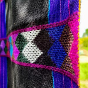 Könnte beinhalten: Nahaufnahme eines Textils mit schwarzem Grund, das ein geometrisches Design in Weiß, Schwarz, Grün und Blau aufweist. Das Design ist von einer leuchtend rosa Bordüre umrandet. Vertikale Streifen in Lila, Blau und Gelb fügen weitere Details hinzu.