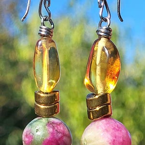 Peut inclure: Boucles d'oreilles pendantes composées d'un crochet argenté, d'une perle ambrée en forme de goutte, de deux perles rectangulaires dorées et d'une pierre ronde rose et verte. Fond vert et bleu flou.