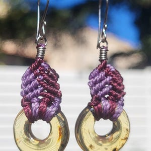 Peut inclure: Paire de boucles d'oreilles faites à la main avec un motif macramé dans les tons violets et bordeaux. Les boucles d'oreilles ont un pendentif circulaire de couleur or et des crochets d'oreilles argentés. Pour oreilles percées.